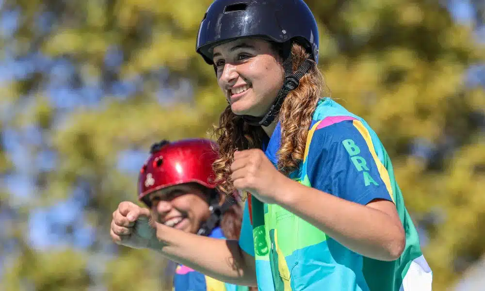 Virgínia Fortes Águas, do skate, comemora fazendo gesto de soquinho com os dois braços. Ela veste uma camiseda estampada colorida e um capacete preto. Rosário 2022 Time Brasil