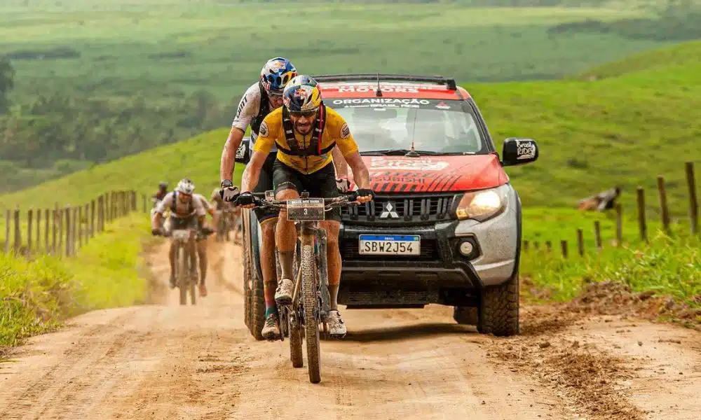 Henrique Avancini pedala em estrada de terra. Ao fundo, outro cicilista e um carro de apoio