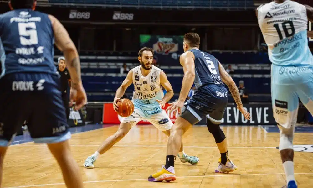 NBB: Jogador do Bauru conduz a bola. Ele veste uniforme branco e azul claro. Ele é marcado por 2 atletas do Brasília, vestindo azul escuro