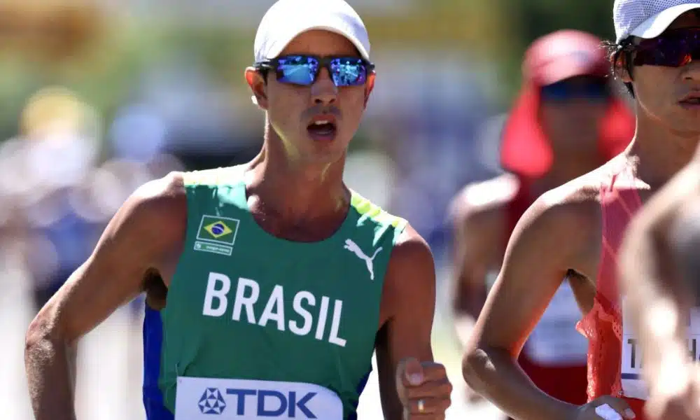 Caio Bonfim marcha vestindo camisa verde da equipe do Brasil de atletismo, boné e óculos de sol
