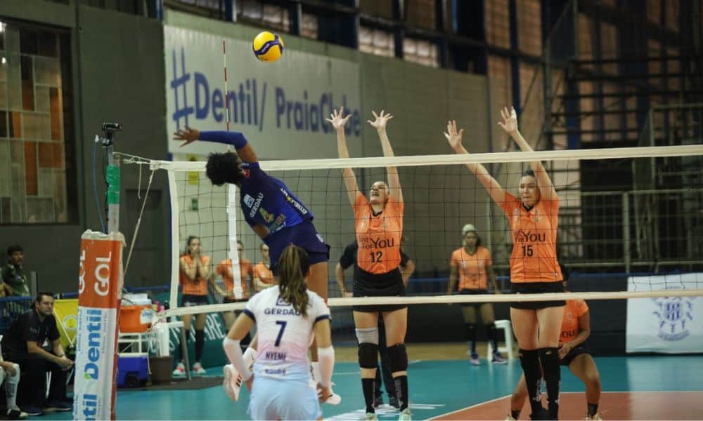 Mineiro de Vôlei - Penã do Minas salta para atacar a bola. Ela é uma mulher negra de uniforme azul escuro. Do outro lado da rede, duas atletas do Vinhedo, sobem no bloqueio