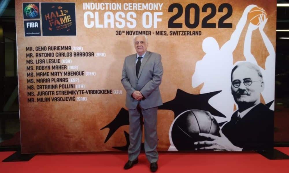 Barbosa, vestindo um terno cinza, posa para foto na frente de banner do hall da fama da FIBA