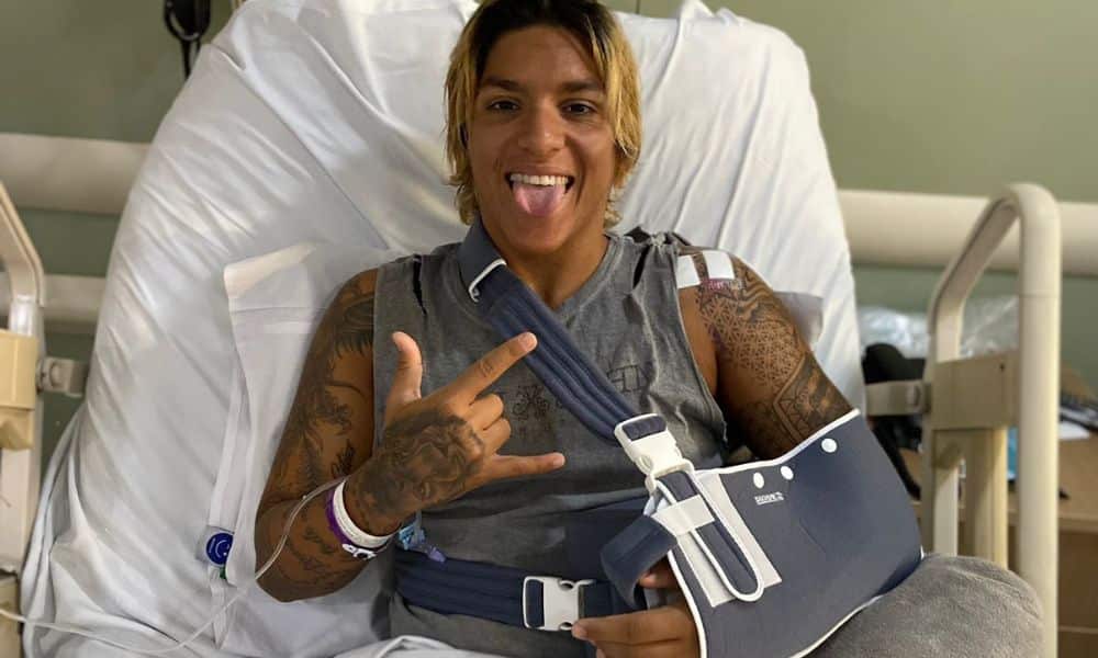Ana Marcela Cunha deitada em cama hospitalar com tipoia no braço esquerdo. Ela faz gesto com a mão e mostra a língua