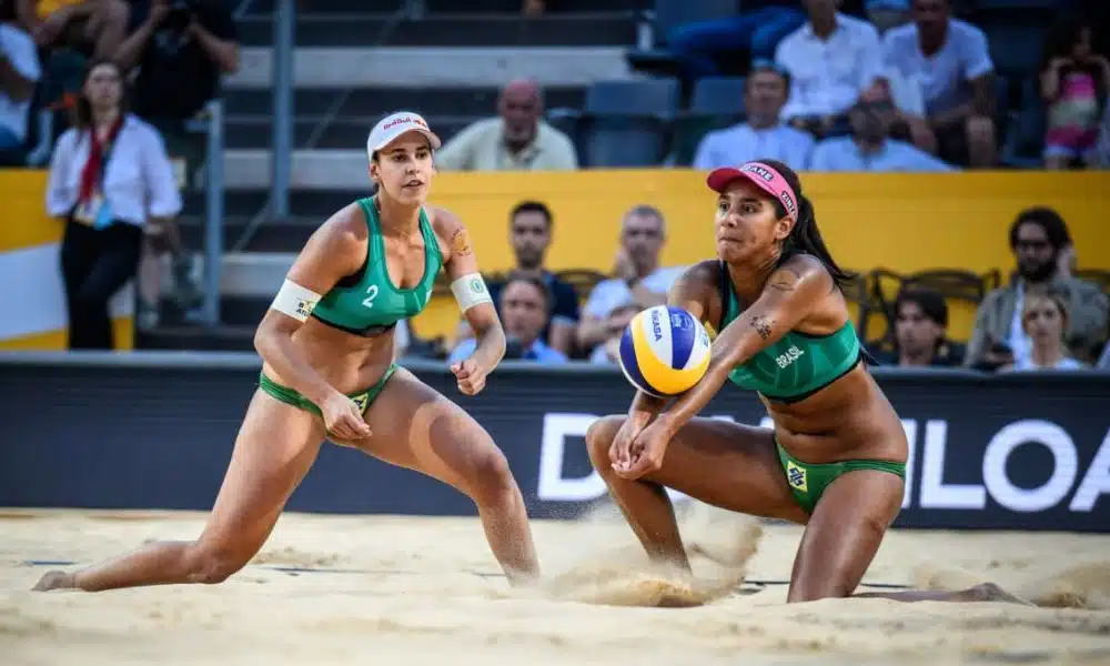 Duda/Ana Patrícia se agacham na areia. Ana faz uma manchete