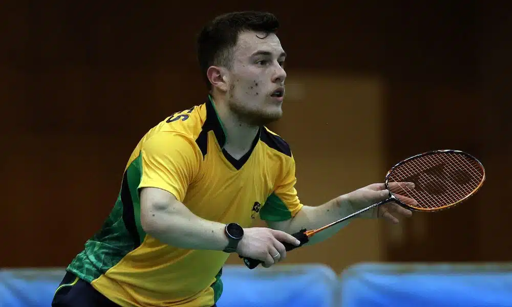 Brasil no parabadminton: Vitor Tavares segura a sua raquete e olha para frente. Ele veste uma camisa amarela do CPB Mundial de parabadminton