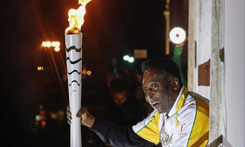 Pelé em uma sacada segurando a tocha branca da Rio-2016 com o fogo aceso. Ele veste uma camisa branca com detalhes em amarelo