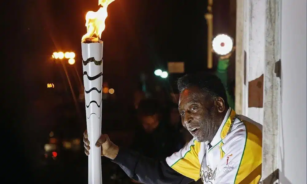Pelé em uma sacada segurando a tocha branca da Rio-2016 com o fogo aceso. Ele veste uma camisa branca com detalhes em amarelo