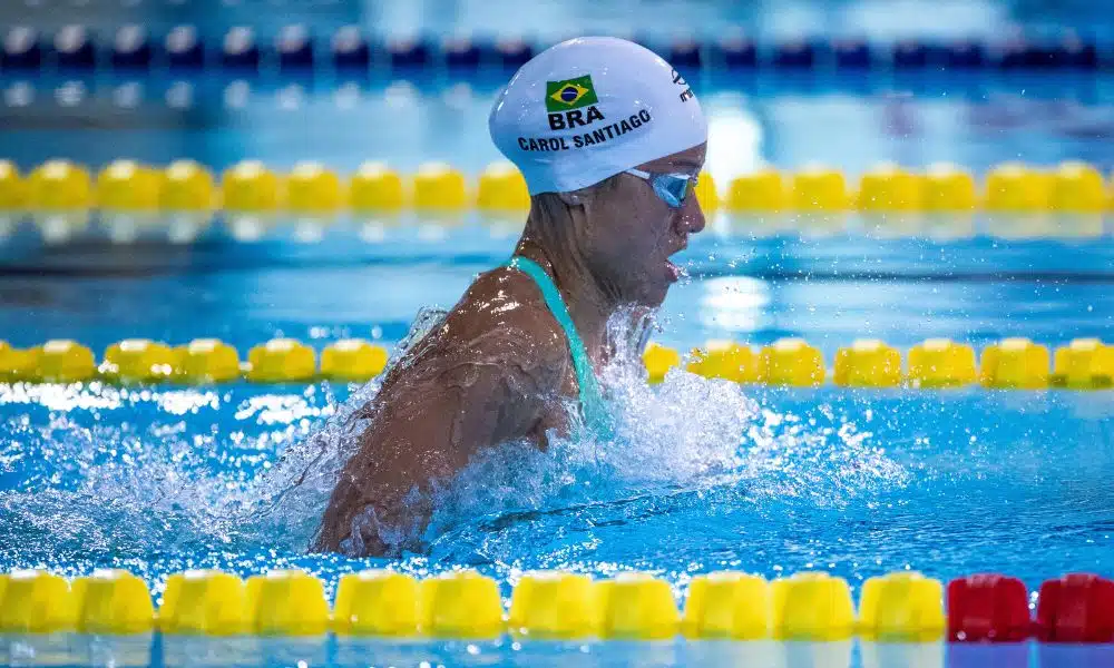 Mas Carol Santiago fazendo o movimento do nado peito. Ela veste maiô azul e touca branca