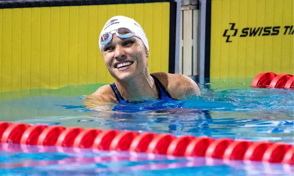 Carol Santiago olha para o lado e sorri na borda da piscina. Ela veste uma touca e óculos de natação brancos / World Series