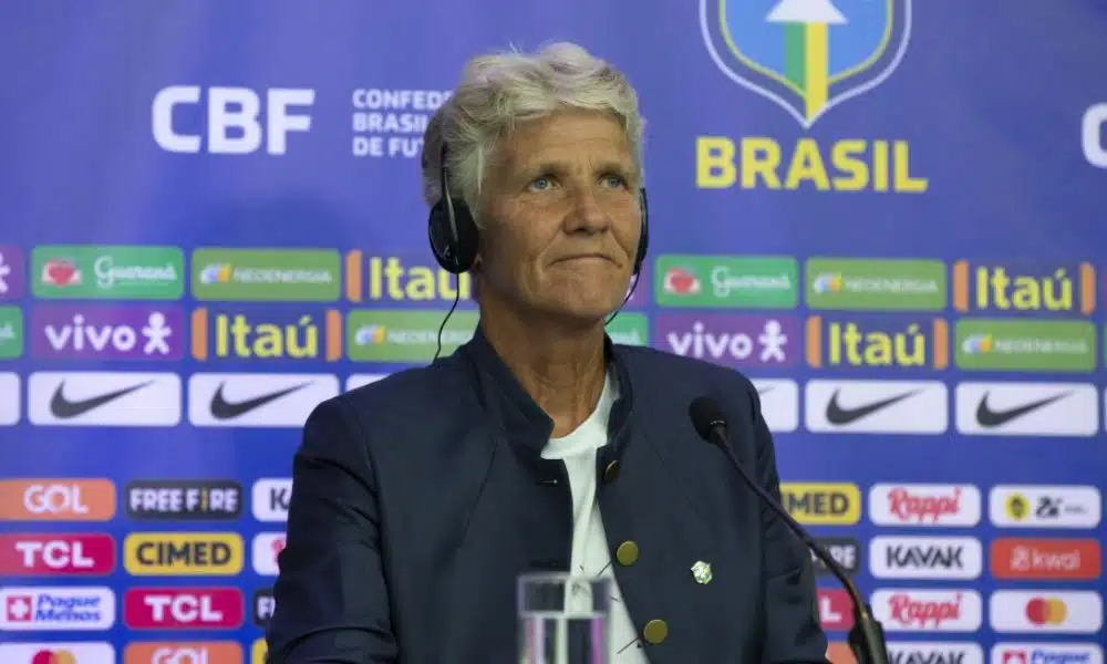 Mas Pia Sundhage na convocação. Ela está sentada em uma mesa e veste uma camiseta branca com um casaco preto por cima. Mas - ao fundo, um mural com patrocinadores da CBF
