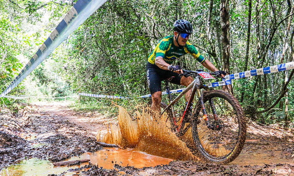 Na image, ciclista do Brasil pedalando e se sujando de lama no Pan-Americano de Mountain Bike.