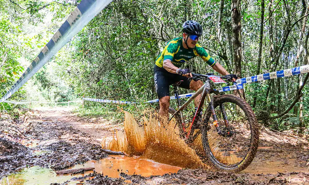 Na image, ciclista do Brasil pedalando e se sujando de lama no Pan-Americano de Mountain Bike.