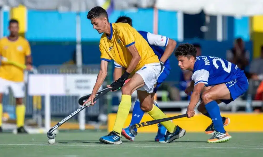 Mas - Jogadores de Brasil e Chile disputam bola em jogo do Pan-Americano júnior de hóquei sobre grama. O brasileiro veste amarelo, enquanto o Chileno tem uniforme azul