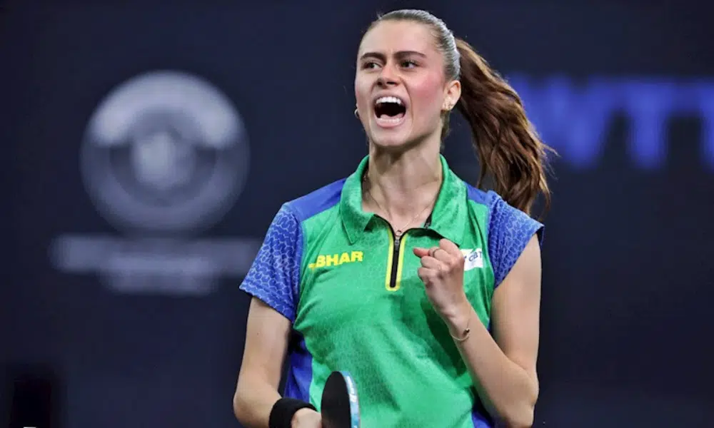 Bruna Takahashi ergue o braço para comemorar um ponto em jogo do Mundial de tênis de mesa. Ela veste uma camiseta verde com mangas azuis e tem o cabelo preso em um rabo de cavalo.