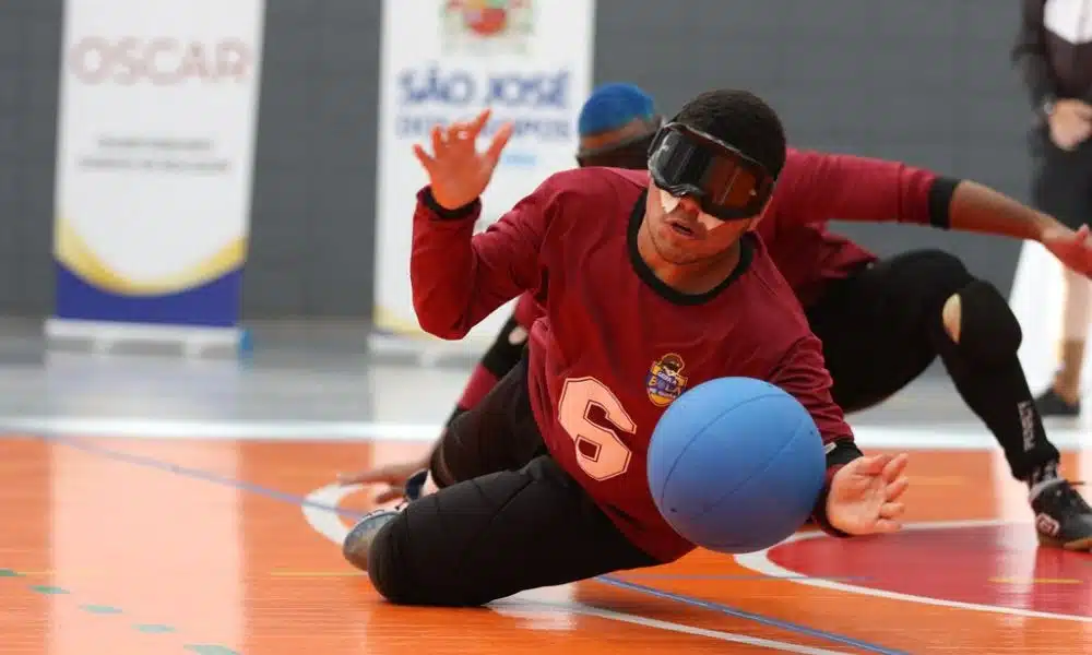 Marco Antônio faz defesa em jogo do Regional Sudeste 2 de goalball. Ele é um jovem negro, veste calça preta e camisa portô e se estica para alcançar a bola