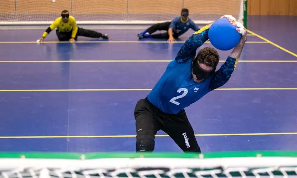 Atletas da seleção masculina de goalball em um treino em São Paulo. No primeiro plano, um jogador segura a bola, enquanto ao fundo, dois atletas estão deitados em posição de defesa