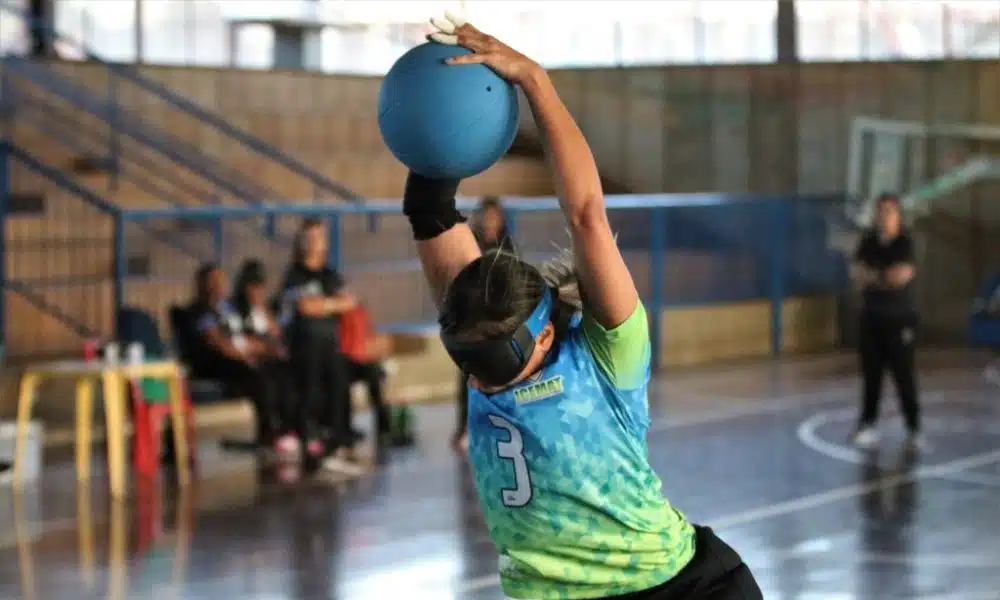 Carol Duarte salta com a bola sobre a cabeça para fazer o arremesso. Foto foi tirada de trás do gol e pega a jogadora de lado, tomando o impulso. Ao fundo, desfocado, o banco de reservas do Cetefe (Foto: Renan Cacioli/ CBDV)