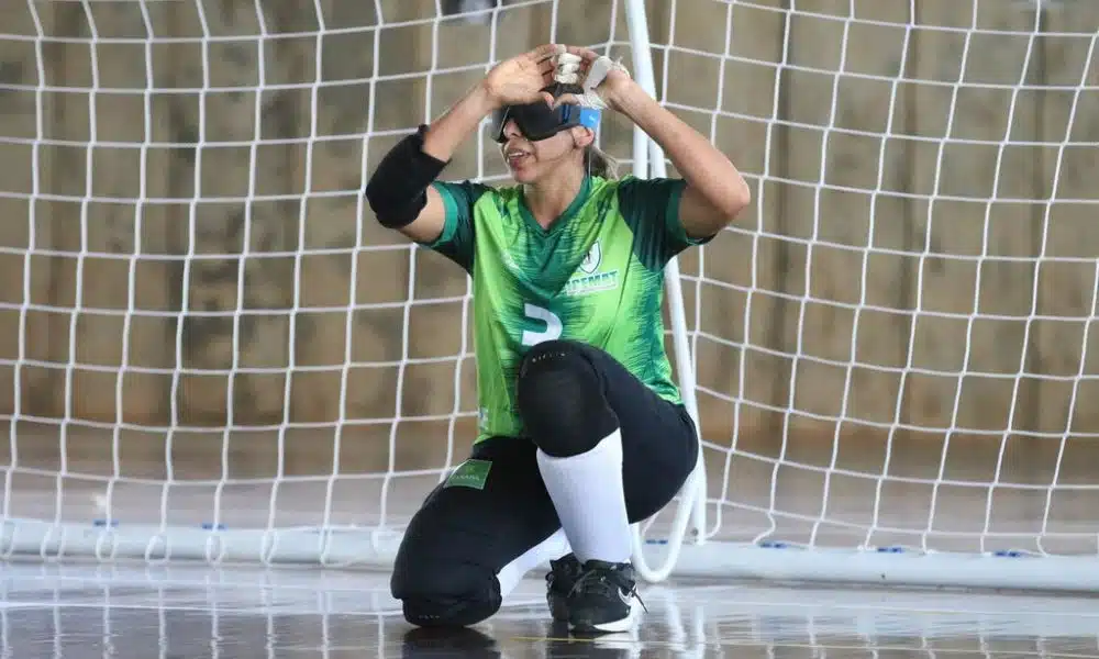 Com o joelho direito apoiado na quadra, Carol Duarte sorri e une as duas mãos à frente do rosto para formar um coração, na comemoração de seu gol na semifinal do Regional Centro-Norte de goalball (Foto: Renan Cacioli/ CBDV)