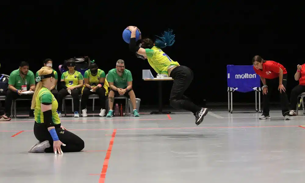 Na imagem, momento em que Dani está saltando com a bola sobre a cabeça para executar o arremesso. Ao seu lado, a pivô Gaby, ajoelhada, aguarda a jogada. Ao fundo, o banco de reserva do Brasil.