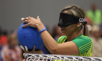 Carol Duarte, do Brasil, está próxima da trave, de costas para a quadra adversária, antes de iniciar seu movimento de arremesso. Ela veste camisa de mangas curtas amarela com detalhes em verde da Seleção e usa esparadrapos nas pontas de alguns dedos das duas mãos.