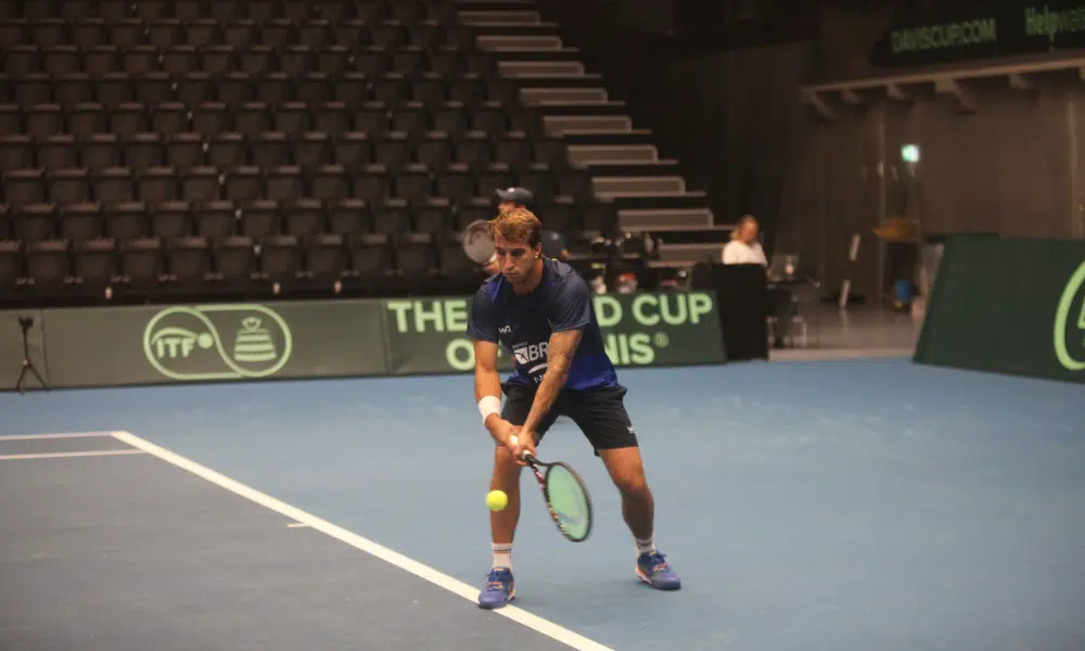 Felipe Meligeni está com raquete na mão, sobre quadra dura, e golpeia a bolinha de tênis com um backhand. Ele e Thiago Wild estão em boa fase e representam o Brasil na Copa Davis