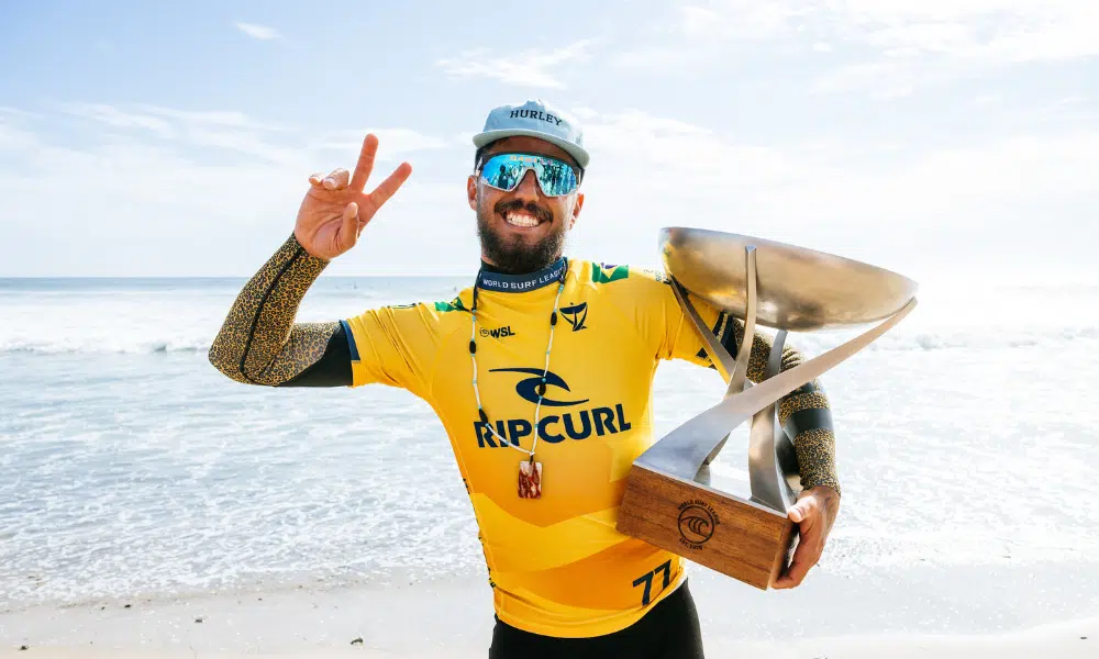 Filipe Toledo sorri, faz gesto com a mão e segura o troféu de campeão do Circuito Mundial de Surfe em frente ao mar