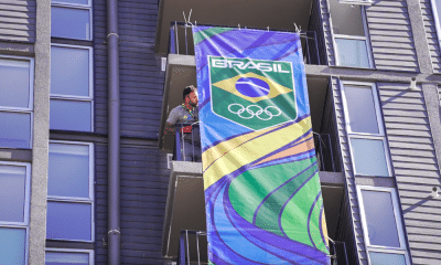 Trabalhador está na sacada de um prédio na Vila Pan-Americana ajustando uma plotagem com a marca do Time Brasil