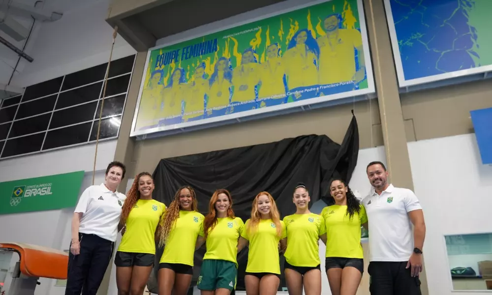 Lorrane Oliveira, Rebeca Andrade, Jade Barbosa, Flávia Saraiva, Julia Soares, Carolyne Pedro e treinadores posam para foto com painel do CT da ginástica no CT Do Time Brasil