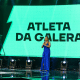 Jornalistas Everaldo Marques (terno cinza) e Karine Alves (vestido azul) seguram o microfone e aparecem no centro da imagem, em cima do palco apresentando o Prêmio Paralímpicos 2022. Ao fundo, há uma tela verde com os dizeres "Atleta da Galera"