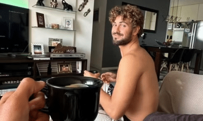 João Chianca, o Chumbinho, aparece de fundo sentado no sofá da sala; em primeiro plano, há um braço esticado com uma caneca