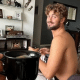 João Chianca, o Chumbinho, aparece de fundo sentado no sofá da sala; em primeiro plano, há um braço esticado com uma caneca