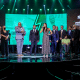 Wilians Araújo e Maria Carol Santiago estão no centro do palco do Prêmio Paralímpicos de 2022, com o troféu de melhores atletas do ano em mãos; ao redor, há diversos dirigentes trajados de terno e gravata