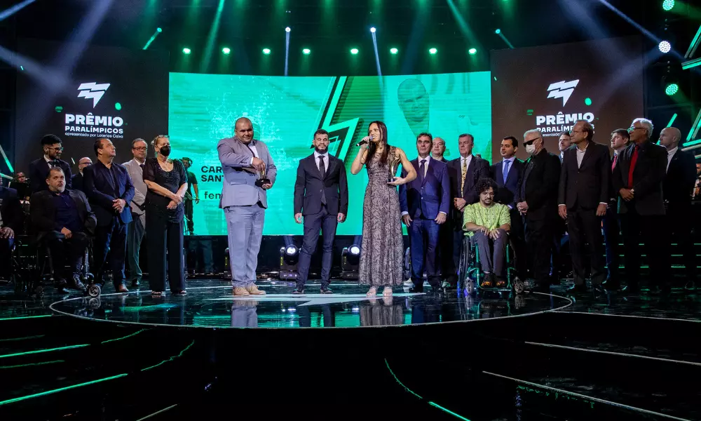 Wilians Araújo e Maria Carol Santiago estão no centro do palco do Prêmio Paralímpicos de 2022, com o troféu de melhores atletas do ano em mãos; ao redor, há diversos dirigentes trajados de terno e gravata