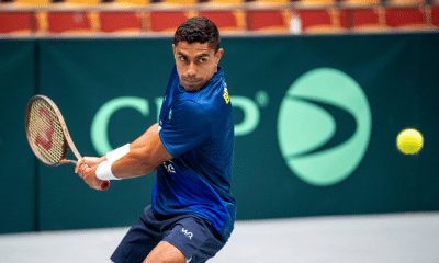 Thiago Monteiro rebate bola; ele está com a raquete nas mãos, de camiseta azul, e faz o movimento de backhand; está em frente ao backdrop da Copa Davis, na Suécia. Brasil também terá Gustavo Heide