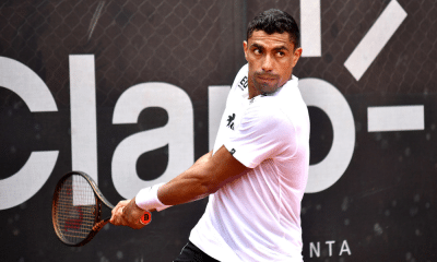 Thiago Monteiro está com a raquete na mão em movimento de ataque em frente ao backdrop do Rio Open; ele enfrentará Carlos Alcaraz na primeira rodada