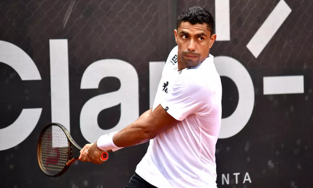 Thiago Monteiro está com a raquete na mão em movimento de ataque em frente ao backdrop do Rio Open; ele enfrentará Carlos Alcaraz na primeira rodada