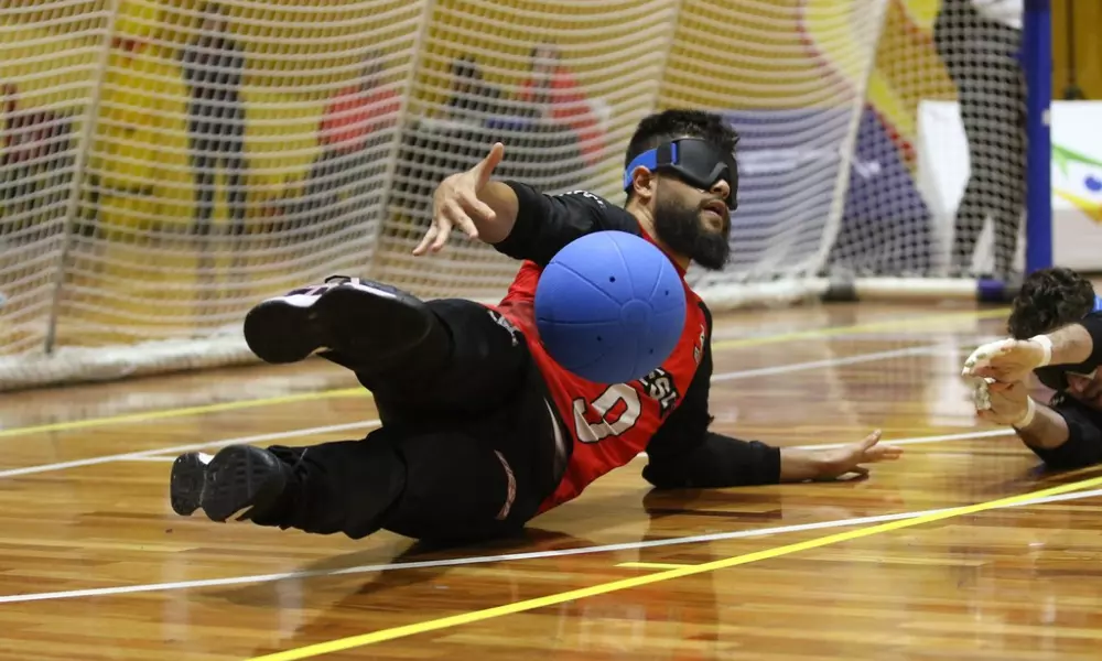 André Dantas cai para o lado esquerdo ao amortecer a bola na defesa com o tronco. Ele veste calça preta e camisa vermelha com mangas longas pretas do Sesi no Regional Sudeste 2 de Goalball