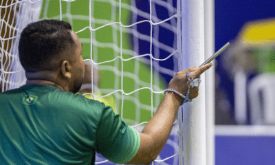 Chamador Cesinha está de costas para a imagem, vestindo camiseta verde de mangas curtas e segura uma barra de metal na mão direita perto de um dos postes da trave, que é branca. Enrolado na mesma mão que segura esta barra há um terço azul. Foto: Alessandra Cabral/CPB.