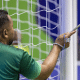 Chamador Cesinha está de costas para a imagem, vestindo camiseta verde de mangas curtas e segura uma barra de metal na mão direita perto de um dos postes da trave, que é branca. Enrolado na mesma mão que segura esta barra há um terço azul. Foto: Alessandra Cabral/CPB.