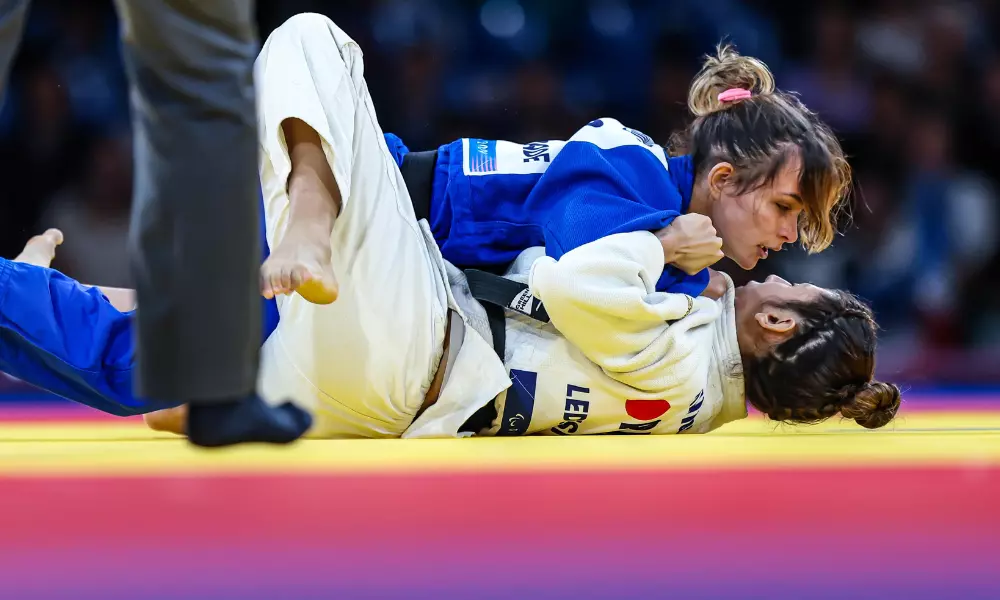 Rosi Andrade, de judogi azul, está no solo imobilizando adversária de branco. O pé da arbitragem, vestindo calça e meia preta, aparece em primeiro plano