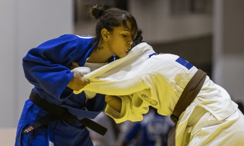 Duas judocas em ação durante uma luta. À esquerda, Brenda Freitas veste um quimono azul e segura firmemente o quimono branco da oponente, cujo rosto está encoberto por seu quimono. Ambas estão usando faixas, sendo a de Brenda preta e a da adversária, marrom. A expressão facial de Brenda demonstra concentração e esforço. O ambiente parece ser um ginásio esportivo, com outras pessoas ao fundo, desfocadas. Foto: Taba Benedicto/CBDV.