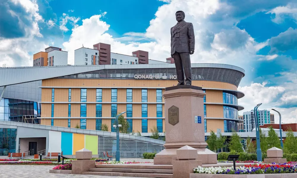 Foto do Palácio Zhaksylyk Ushkempirov, localizado em Astana, que possui uma estátua de um homem vestindo terno à frente, sobre um pedestal. Ao fundo, o edifício é moderno e tem fachada em tons de bege e azul, com janelas espelhadas. O céu está azul com algumas nuvens brancas, e há canteiros de flores coloridas na área em frente à estátua. Ao redor, há postes de iluminação modernos e algumas árvores. Foto: Divulgação.