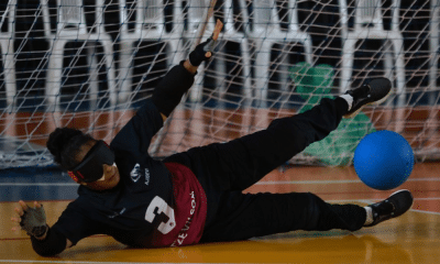 Vestindo uniforme preto com o número 3, venda preta e luvas sem dedos, Adria está deitada de lado em uma quadra coberta, bloqueando a bola azul que rola nas proximidades. Atrás dela, há uma rede. O fundo mostra cadeiras brancas e alguns objetos verdes atrás da rede. A linguagem corporal da atleta indica foco e esforço enquanto ela estica os braços e as pernas. Foto: Itamar Aguiar/ CBDV