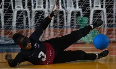 Vestindo uniforme preto com o número 3, venda preta e luvas sem dedos, Adria está deitada de lado em uma quadra coberta, bloqueando a bola azul que rola nas proximidades. Atrás dela, há uma rede. O fundo mostra cadeiras brancas e alguns objetos verdes atrás da rede. A linguagem corporal da atleta indica foco e esforço enquanto ela estica os braços e as pernas. Foto: Itamar Aguiar/ CBDV