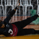 Vestindo uniforme preto com o número 3, venda preta e luvas sem dedos, Adria está deitada de lado em uma quadra coberta, bloqueando a bola azul que rola nas proximidades. Atrás dela, há uma rede. O fundo mostra cadeiras brancas e alguns objetos verdes atrás da rede. A linguagem corporal da atleta indica foco e esforço enquanto ela estica os braços e as pernas. Foto: Itamar Aguiar/ CBDV