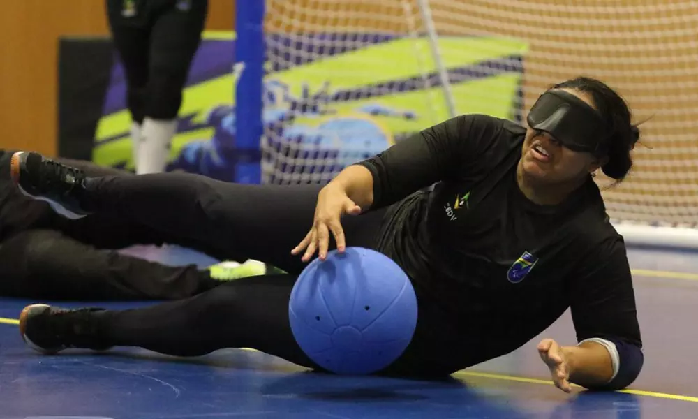 Jéssica está deitada no chão de uma quadra esportiva, usando uma venda nos olhos. Ela veste o uniforme preto da CBDV e aparece após interceptar uma bola azul que está próxima a ela. Ao fundo, há uma rede de gol e parte de uma parede com cores vivas. Crédito: Renan Cacioli/ CBDV.