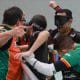 Na imagem, Grupo da AGAFUC comemora na banda lateral. Dois atletas, Gabriel e Nonato, usam máscaras pretas para os olhos e protetores bucais. Os jogadores se abraçam e parecem muito felizes. De camiseta laranja e verde, o técnico Rafael Astrada estende a mão, enquanto o goleiro Luan, de uniforme verde, está curvado no abraço coletivo. O fundo é um ginásio esportivo coberto com uma parede de tijolos cinza.