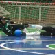 Quadra esportiva com duas jogadoras do ICEMAT deitadas no chão, próximas à linha do gol. Ambas usam vendas nos olhos e uniforme preto com listras verdes. Uma delas, com o número 5, está sentada com uma das mãos apoiada no chão e a outra perna dobrada, enquanto a bola azul está encostada em sua perna. A outra jogadora está totalmente deitada, com os braços esticados para frente, como se estivesse tentando defender a bola. Ao fundo, há uma trave de gol e arquibancadas vazias com cadeiras verdes