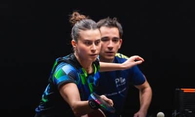 Na imagem, Bruna Takahashi rebatendo a bolinha com sua raquete e Hugo Calderano observando de perto.