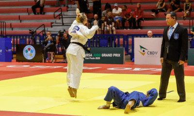 Rosi Andrade, de quimono branco, está saltando com ambos os pés para fora do chão, aparentemente comemorando. No chão, uma oponente de quimono azul está deitada de costas